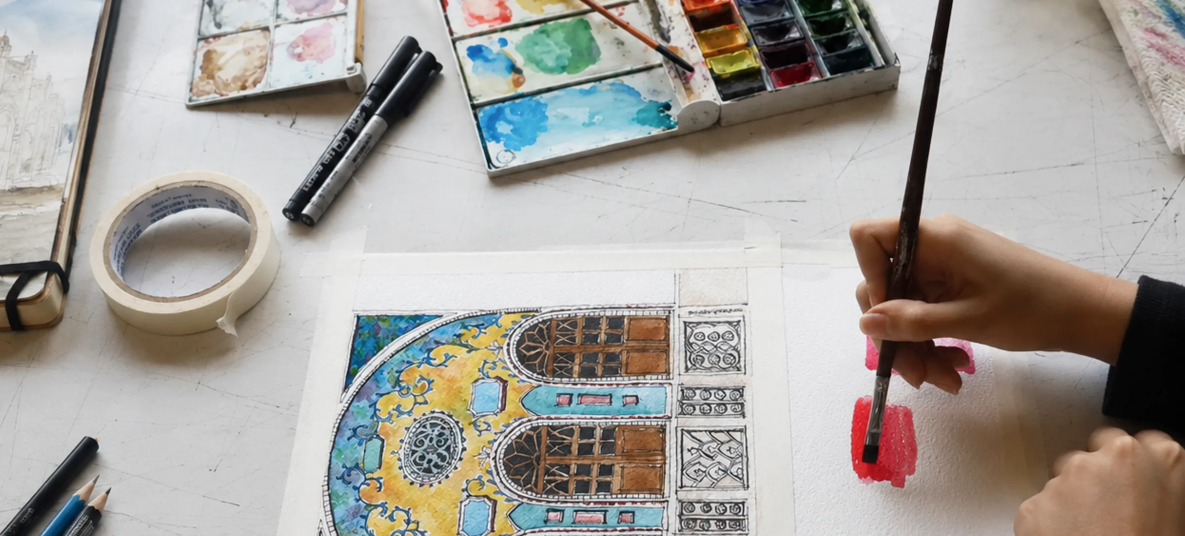 Watercolor paints, markers, and a hand painting an architectural illustration on a studio desk